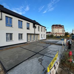 Firma Usługowo-Handlowa Andrzej Mydło - Nowoczesna posesja z ułożoną szarą kostką brukową, widok na budynek mieszkalny i fragment ogrodzenia z żółtą taśmą. Błękitne niebo w tle.