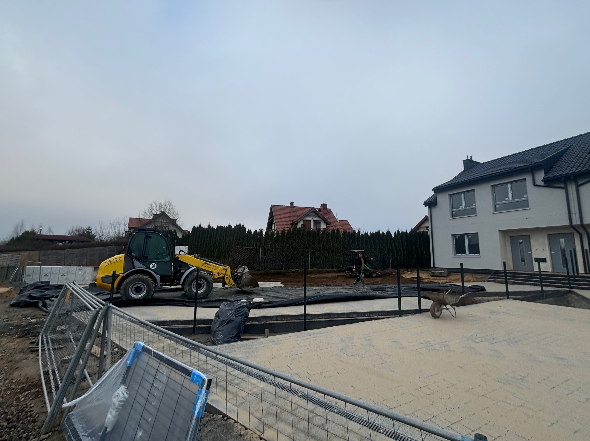 Żółta ładowarka teleskopowa na placu budowy z częściowo ułożoną kostką. W tle domy i pracownik z taczką. Widoczne tymczasowe ogrodzenie budowlane.