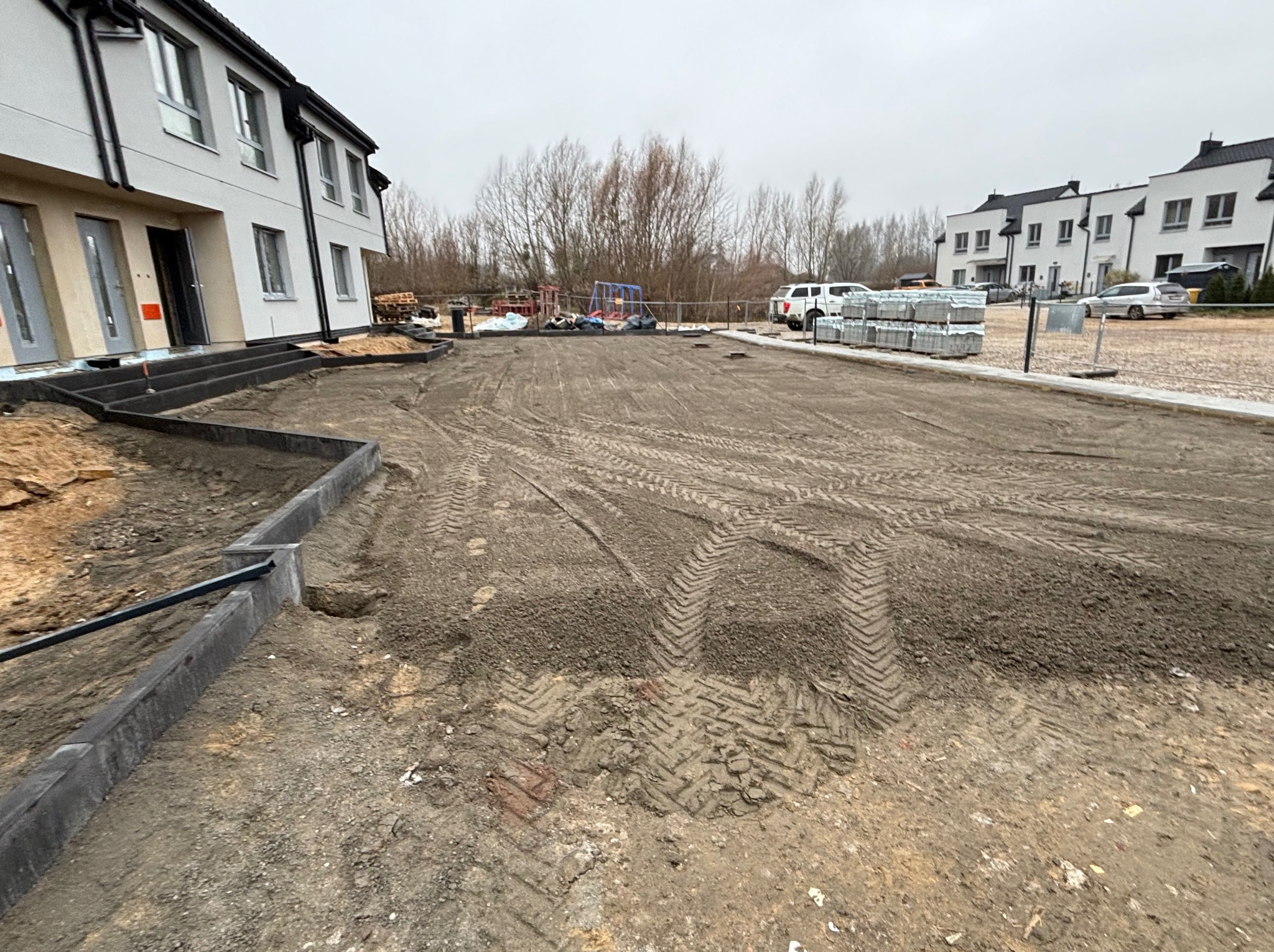 Przygotowany teren pod budowę z obrzeżami i śladami opon, w tle nowe budynki mieszkalne i palety z kostką. Pochmurny dzień.