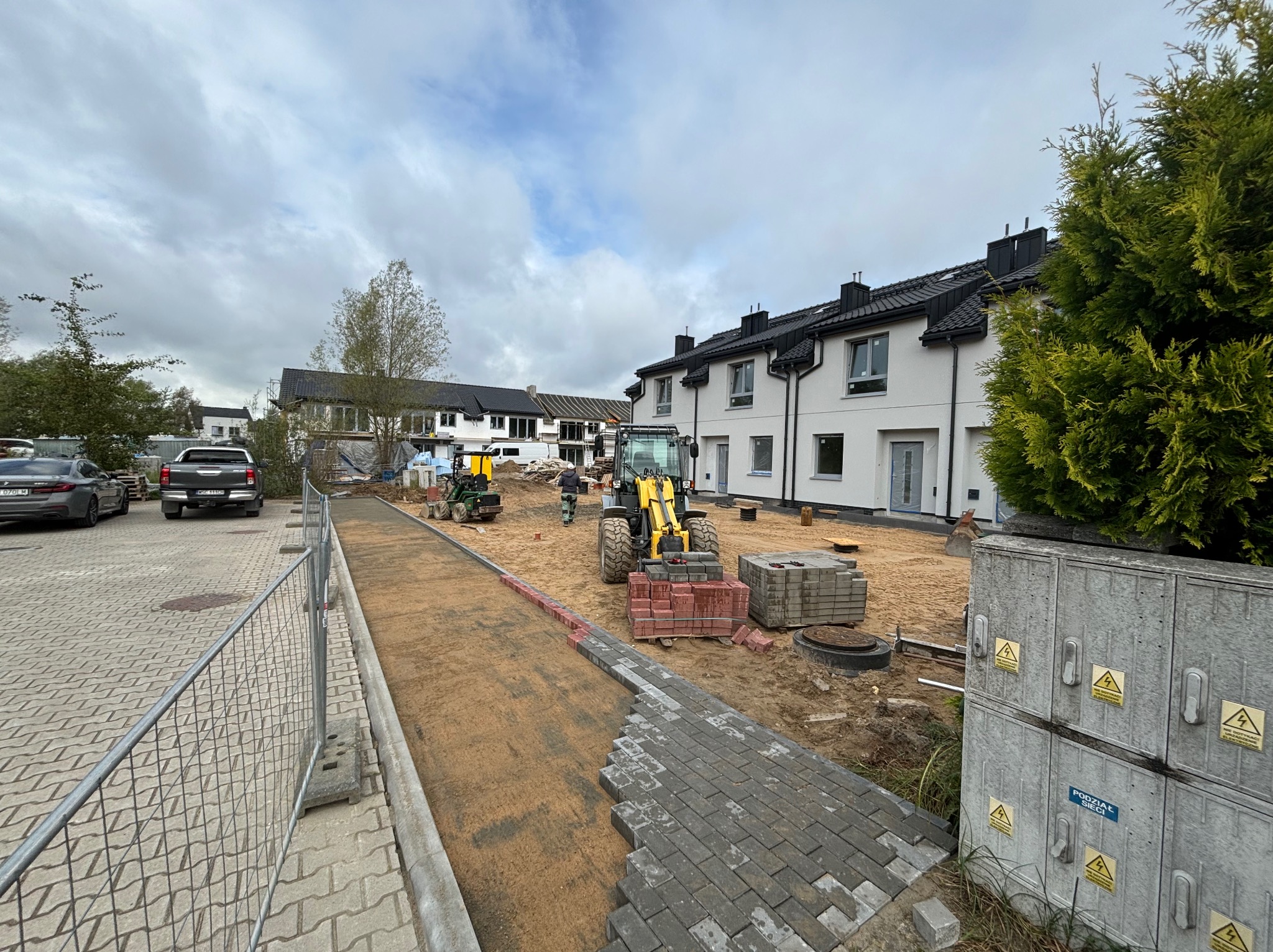 Fragment układanej szarej kostki brukowej na podsypce piaskowej, w tle budynki mieszkalne i maszyny budowlane. Widoczne palety z kostką i tymczasowe ogrodzenie.