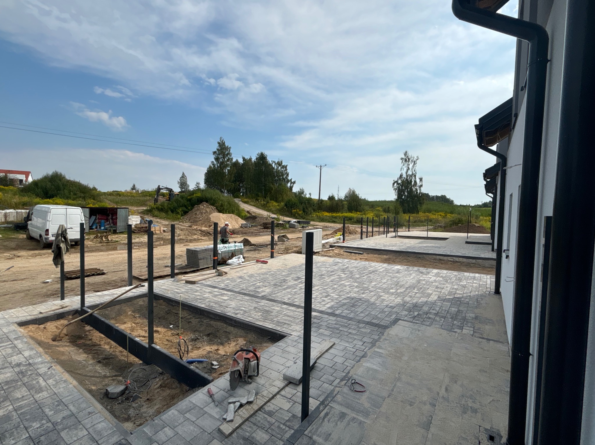 Fragment posesji w trakcie prac: ułożona kostka brukowa, przygotowane fundamenty pod ogrodzenie panelowe, widoczny sprzęt budowlany i teren w trakcie niwelacji.