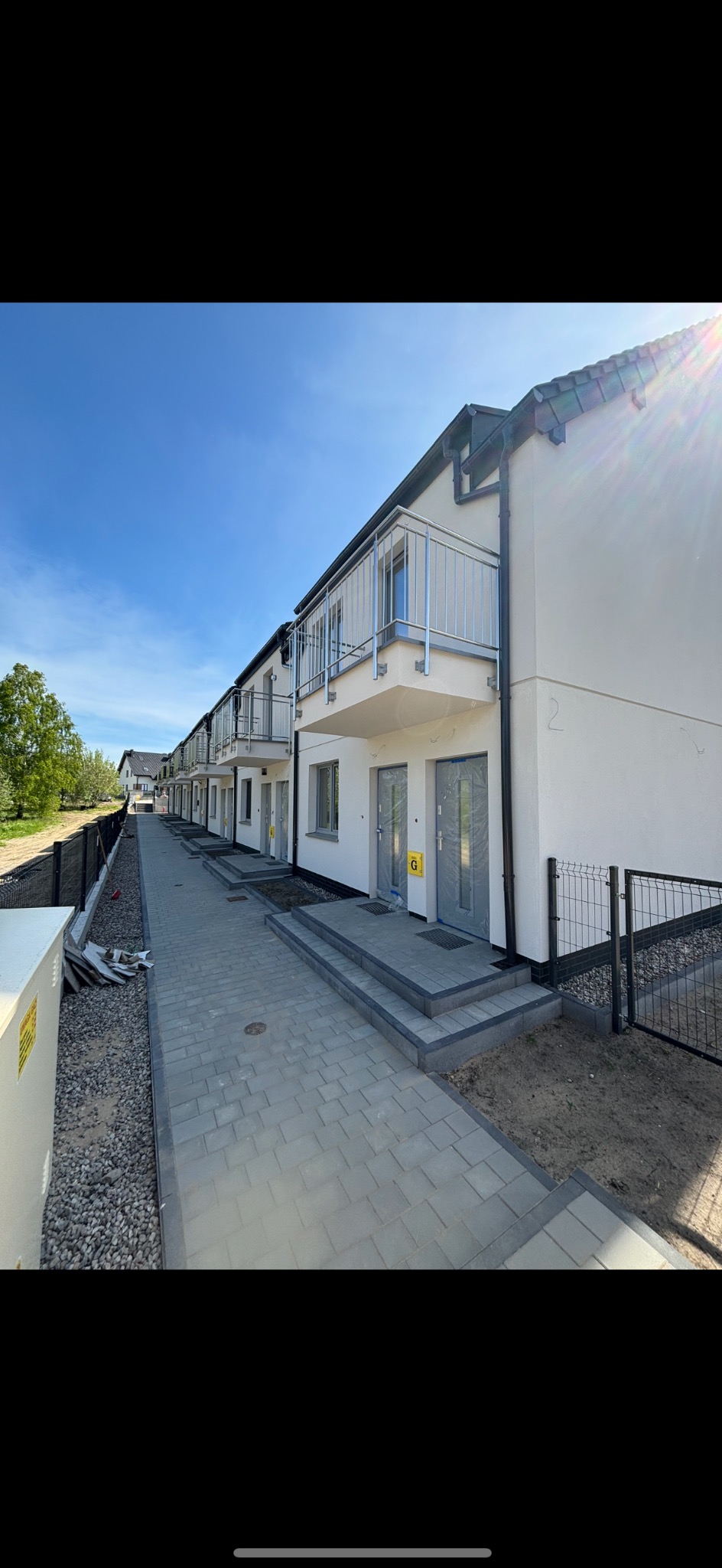 Szara kostka brukowa prowadzi do szeregu nowych domów z balkonami i ogrodzeniem z siatki. Słoneczny dzień.