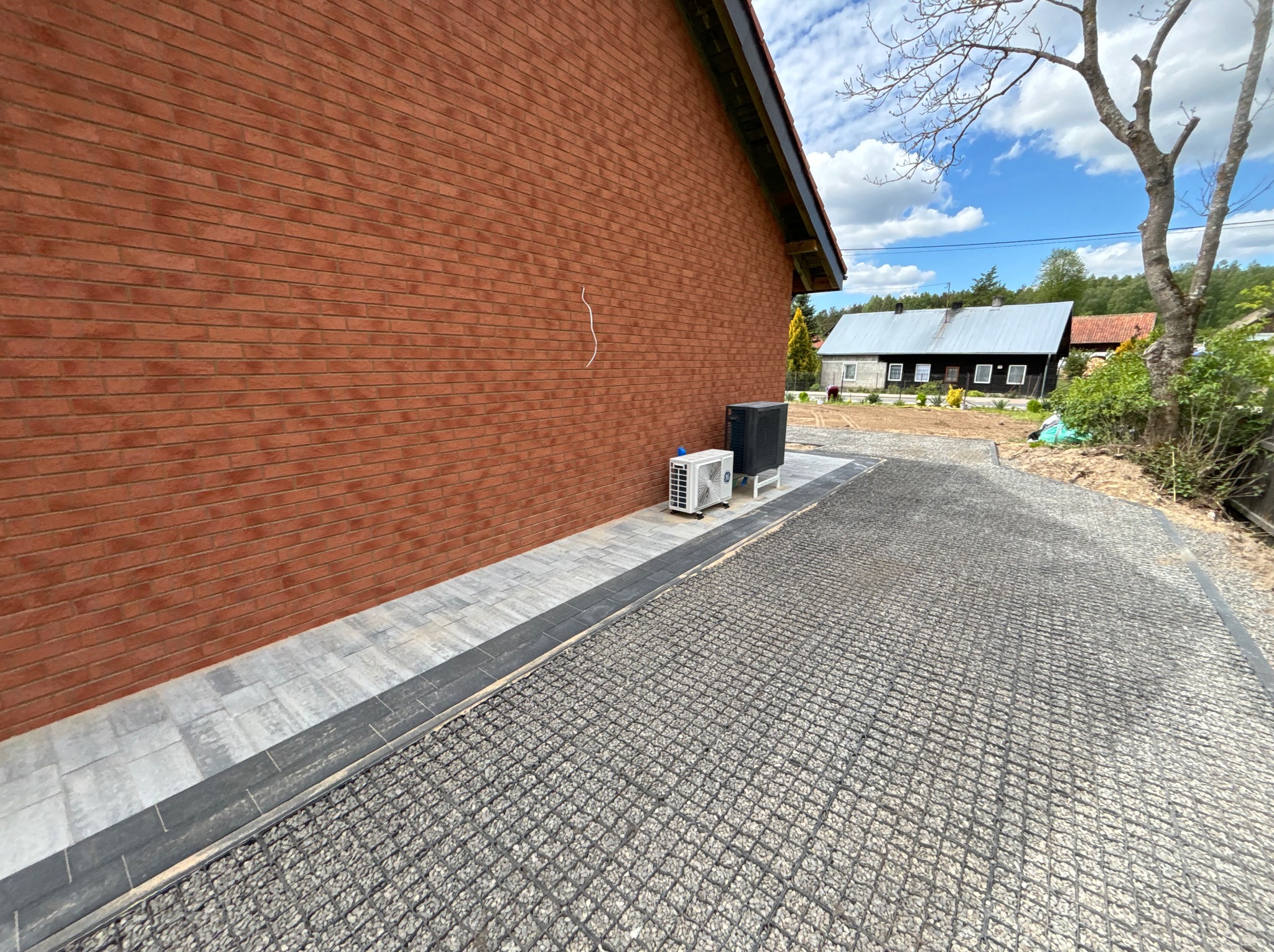Utwardzona droga z kratki trawnikowej i obrzeżem z kostki, obok budynku z cegły. Widoczne klimatyzatory i budynek w tle.