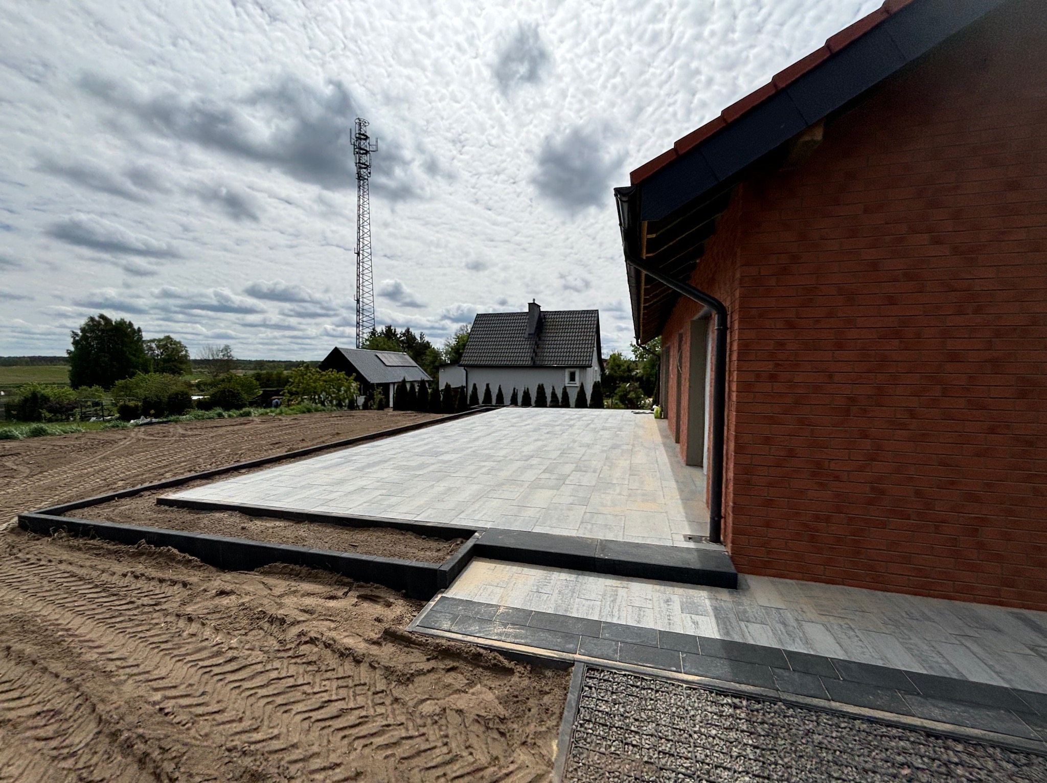 Ułożona kostka brukowa na posesji z widokiem na dom i wieżę telekomunikacyjną w tle. Czarna obwódka kostki. Ślady opon na ziemi.