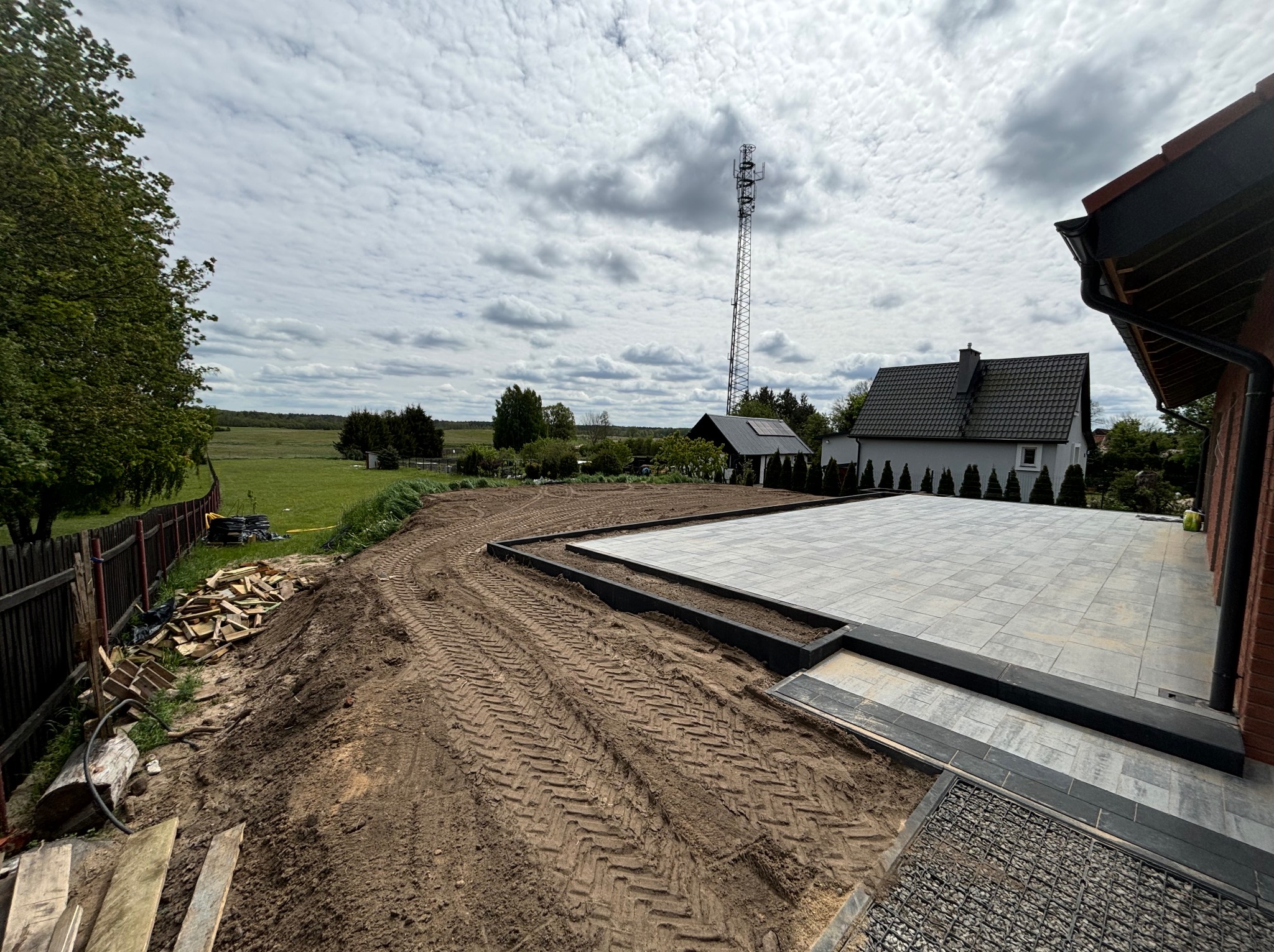 Ułożona kostka brukowa na tarasie domu jednorodzinnego, widoczne ślady opon na ziemi i przygotowany teren pod dalsze prace brukarskie. W tle dom i wieża telekomunikacyjna.
