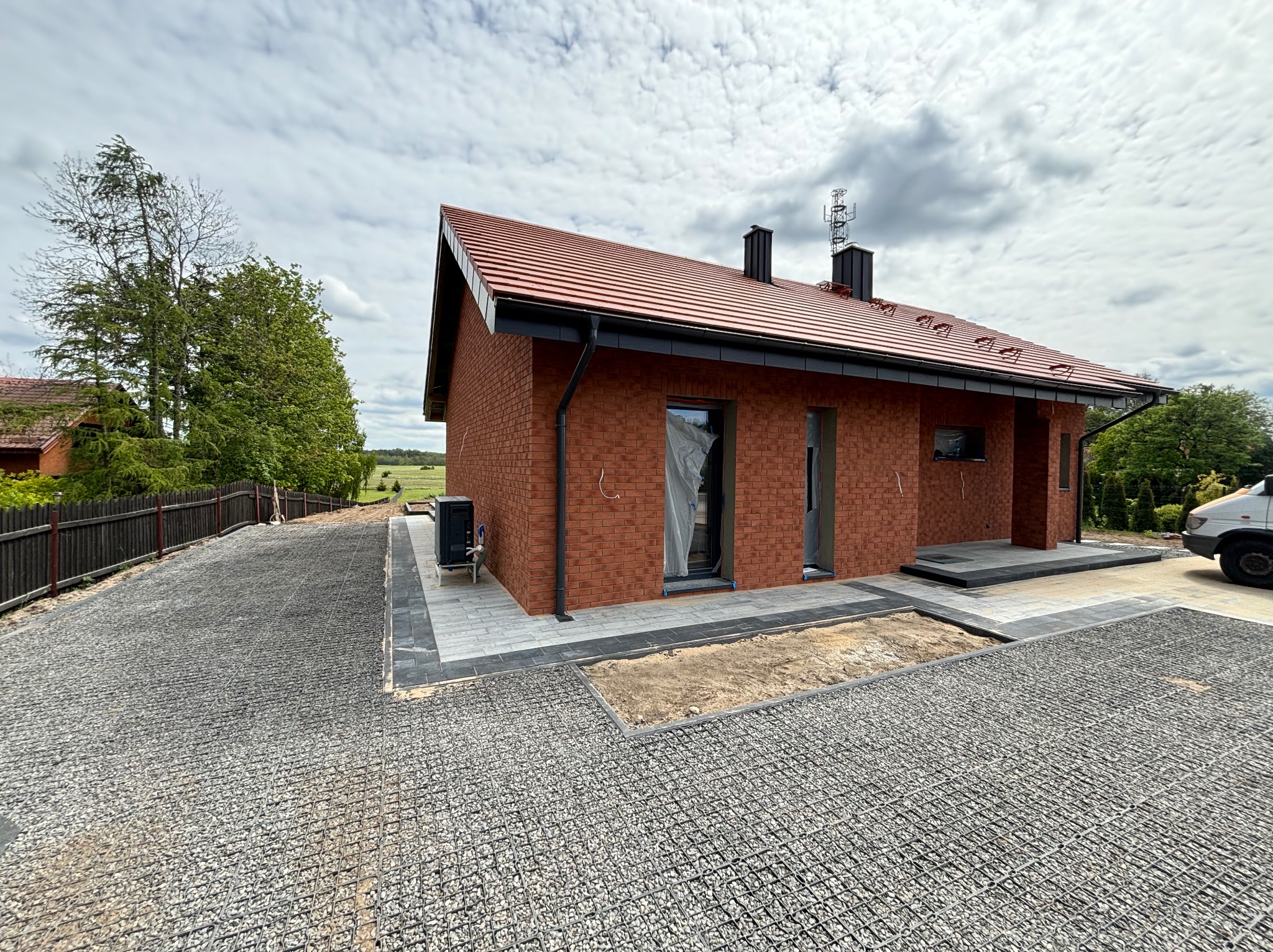 Nowoczesny dom z czerwonej cegły z wykończonym podjazdem z kruszywa w siatce i kostki. Widoczne elementy wykończeniowe i fragment ogrodu.