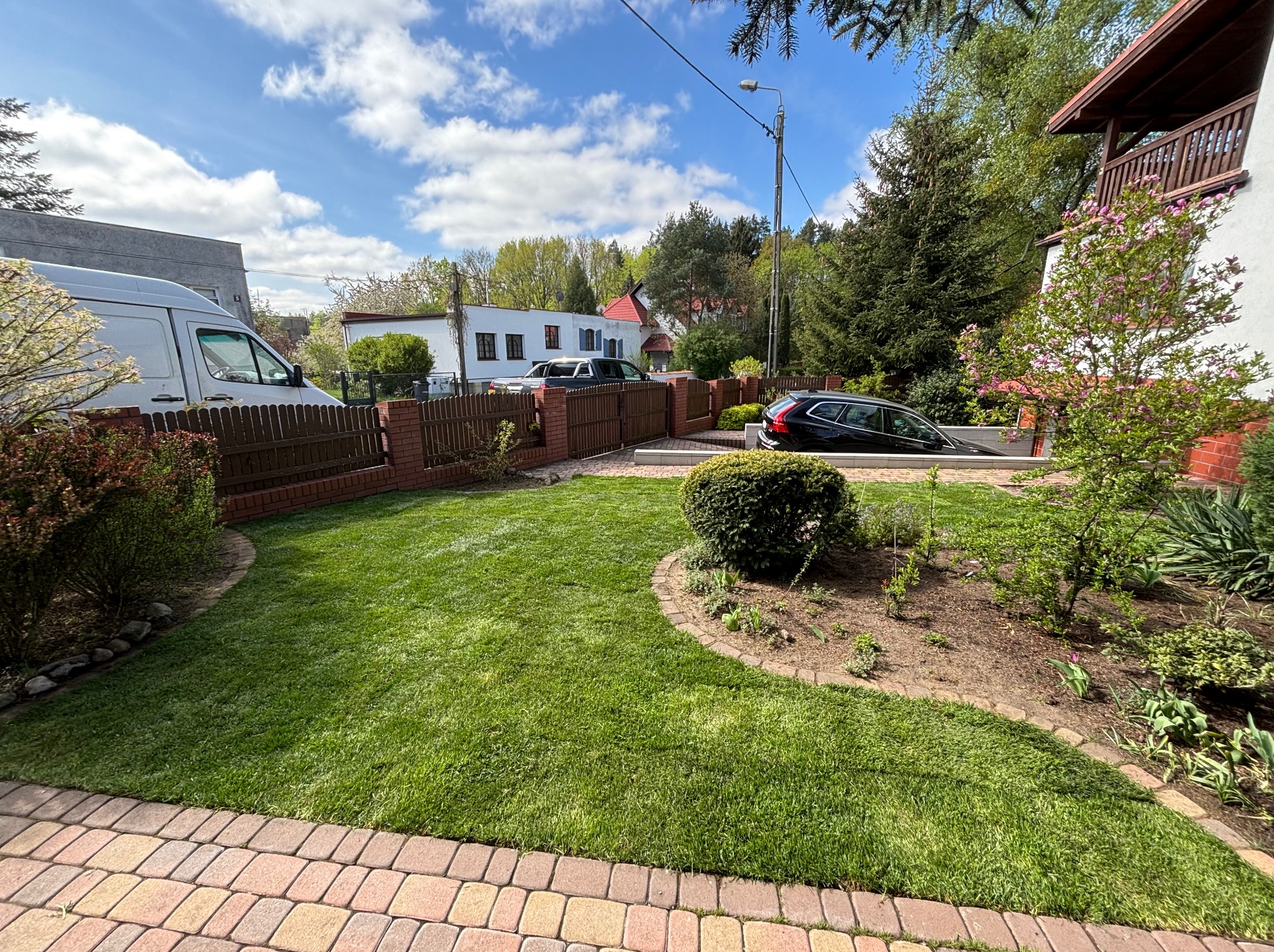Zadbany trawnik z obrzeżem z kostki, ogrodzenie z cegły i drewna, krzewy ozdobne, dom z balkonem w tle, słoneczny dzień.