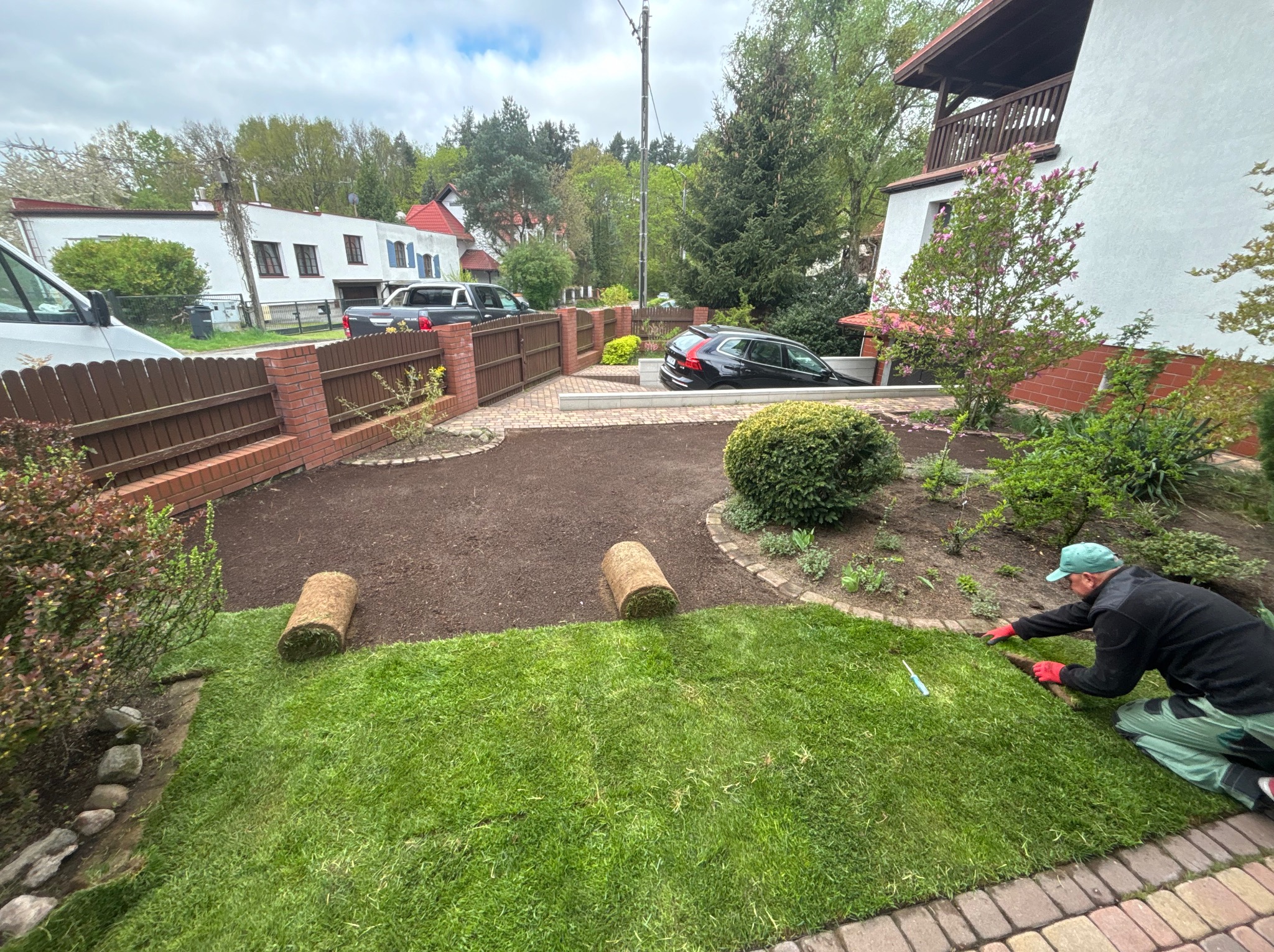 Układanie trawy z rolki w ogrodzie. Widoczny fragment trawnika, rolki trawy i osoba w trakcie pracy, w tle dom i ogrodzenie z cegły.