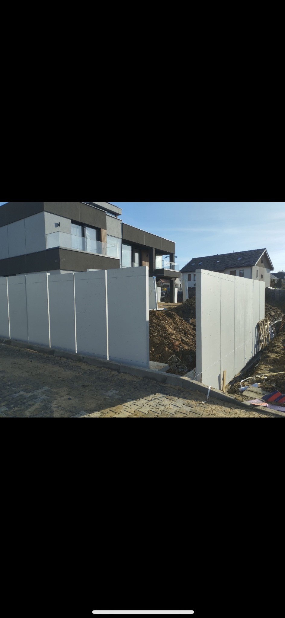 Montaż prefabrykowanych elementów ogrodzenia betonowego na posesji z nowoczesnym domem. Widoczne wykopane fundamenty i przygotowany teren pod dalsze prace.