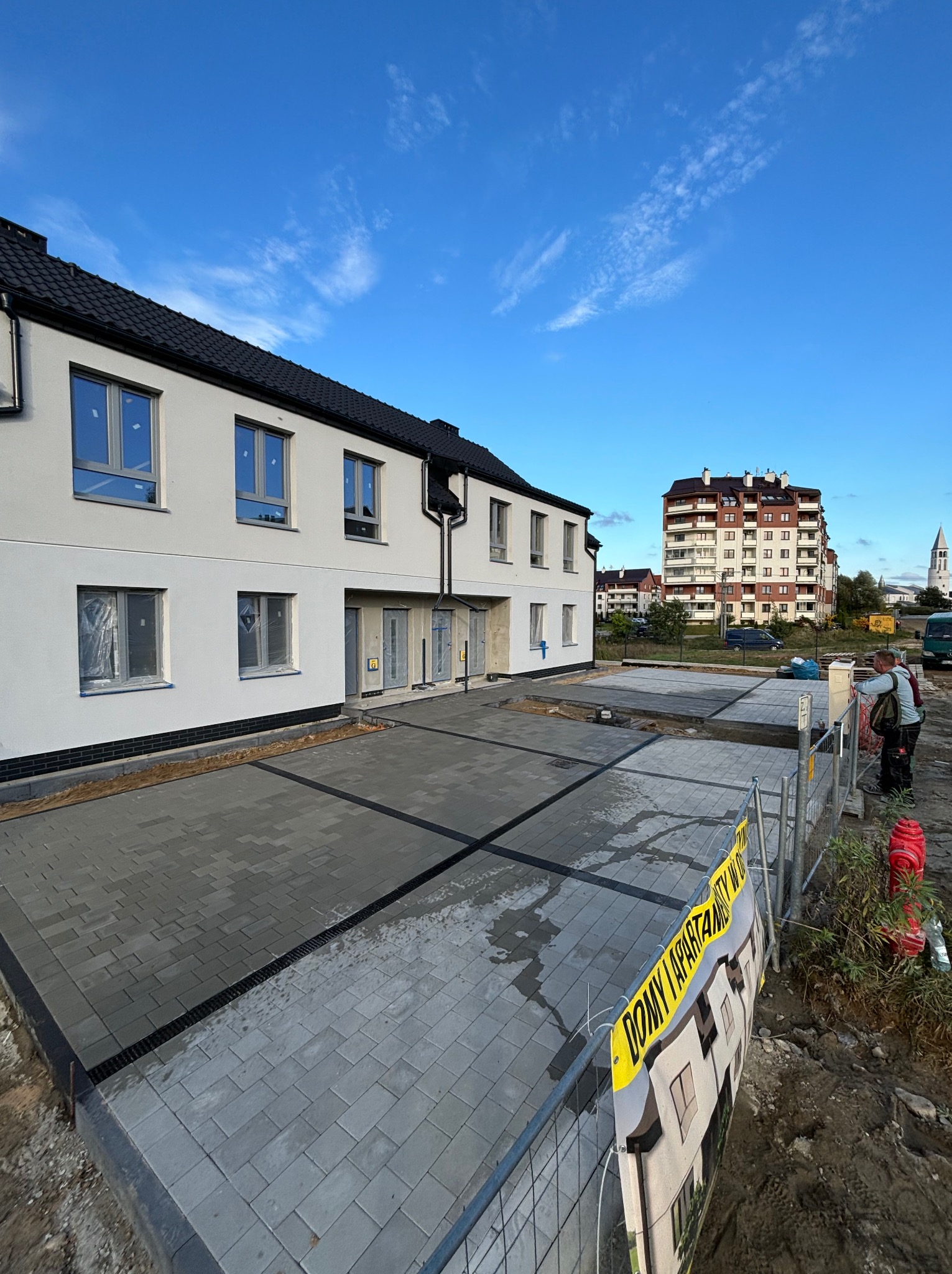 Nowoczesna posesja z ułożoną szarą kostką brukową, widok na budynek mieszkalny i fragment ogrodzenia z żółtą taśmą. Błękitne niebo w tle.