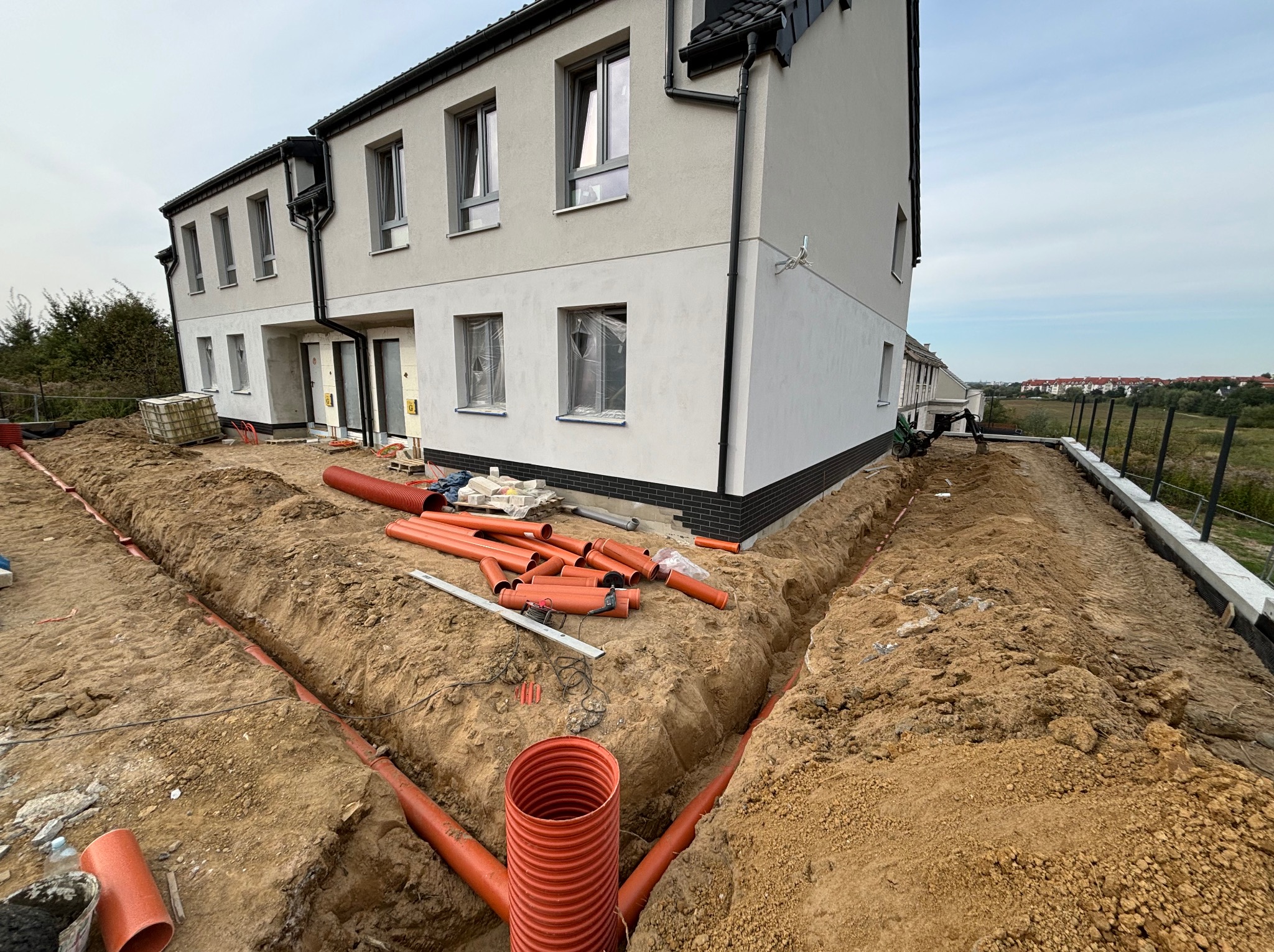 Instalacja rur drenarskich wokół nowego domu. Wykopane rowy, pomarańczowe rury w ziemi, narzędzia pracy. Widok na budynek i okolicę. Prace ziemne w toku.