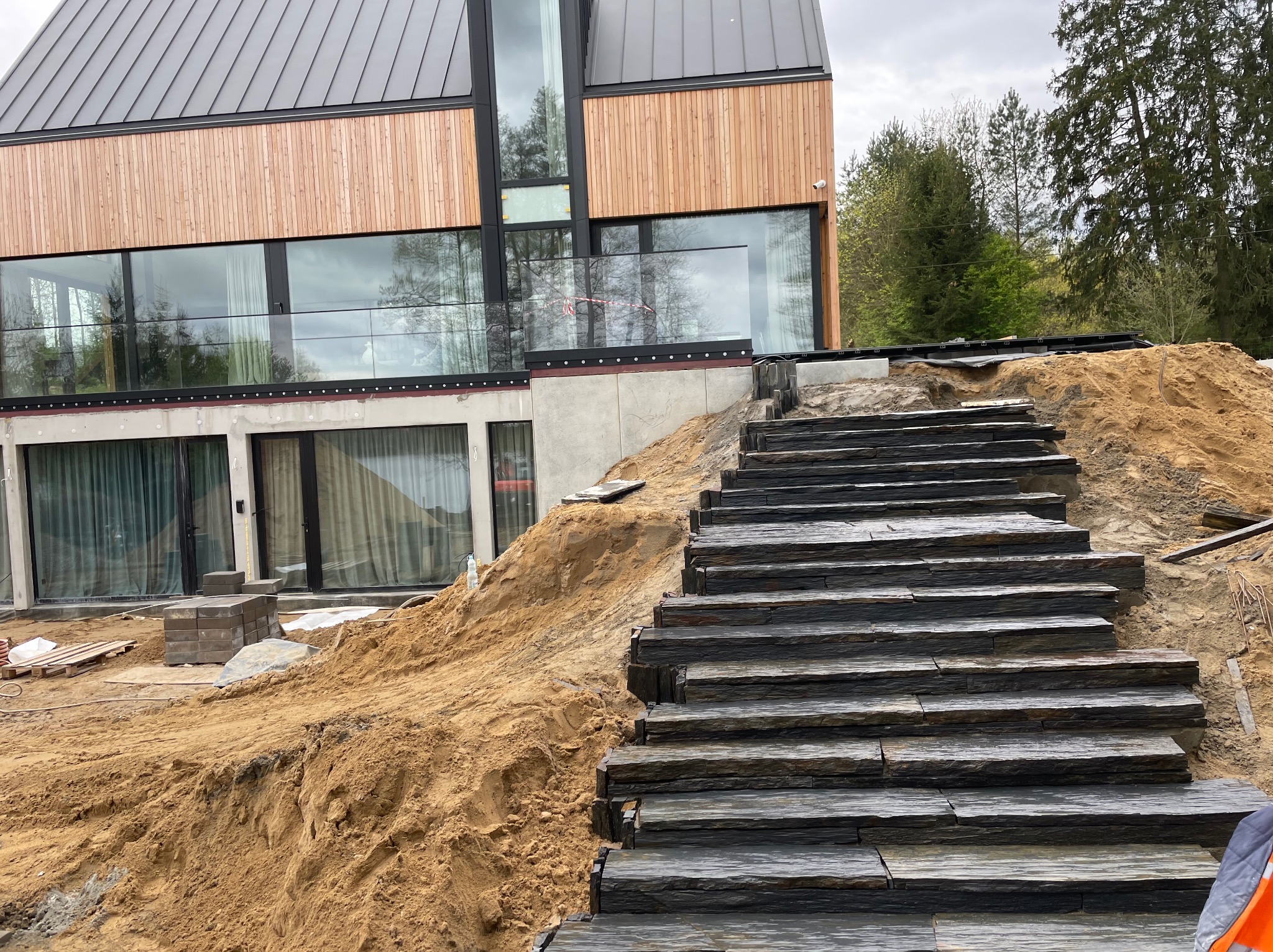 Nowoczesny dom z kamiennymi schodami w trakcie budowy. Widoczne ułożone stopnie z ciemnego kamienia i przygotowany teren pod dalsze prace brukarskie.