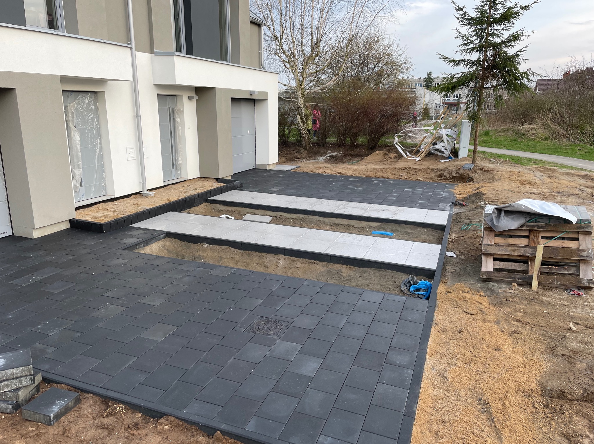 Częściowo ułożona, grafitowa kostka brukowa przed nowoczesnym domem w trakcie realizacji. Widoczne przygotowane podłoże i paleta. Ujęcie z góry.