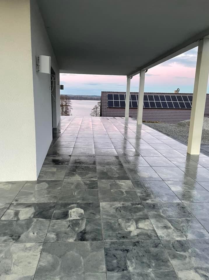 Zadaszony taras z szarych, wzorzystych płyt. W tle jezioro i dom z panelami słonecznymi. Widok z perspektywy niskiego kąta, podkreślający fakturę płytek.