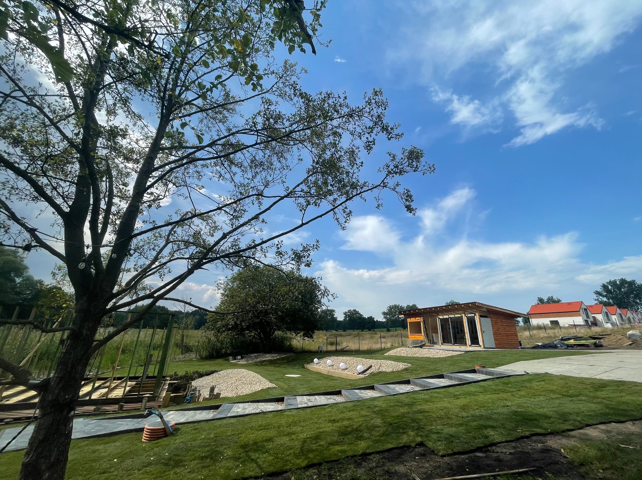 Aranżacja ogrodu w Olsztynie: ścieżka z kostki, trawnik, rabaty z kamyków i oświetleniem. W tle domek letniskowy i budowa ogrodzenia.