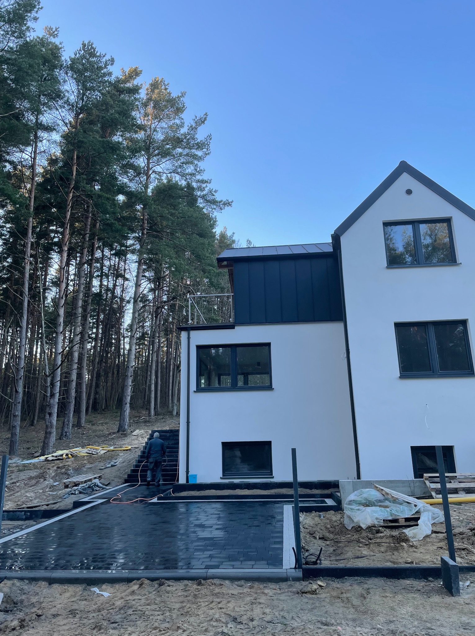 Ciemna kostka brukowa ułożona przed nowoczesnym domem z białymi ścianami i ciemnymi oknami. W tle las. Osoba wchodzi po schodach z kostki.