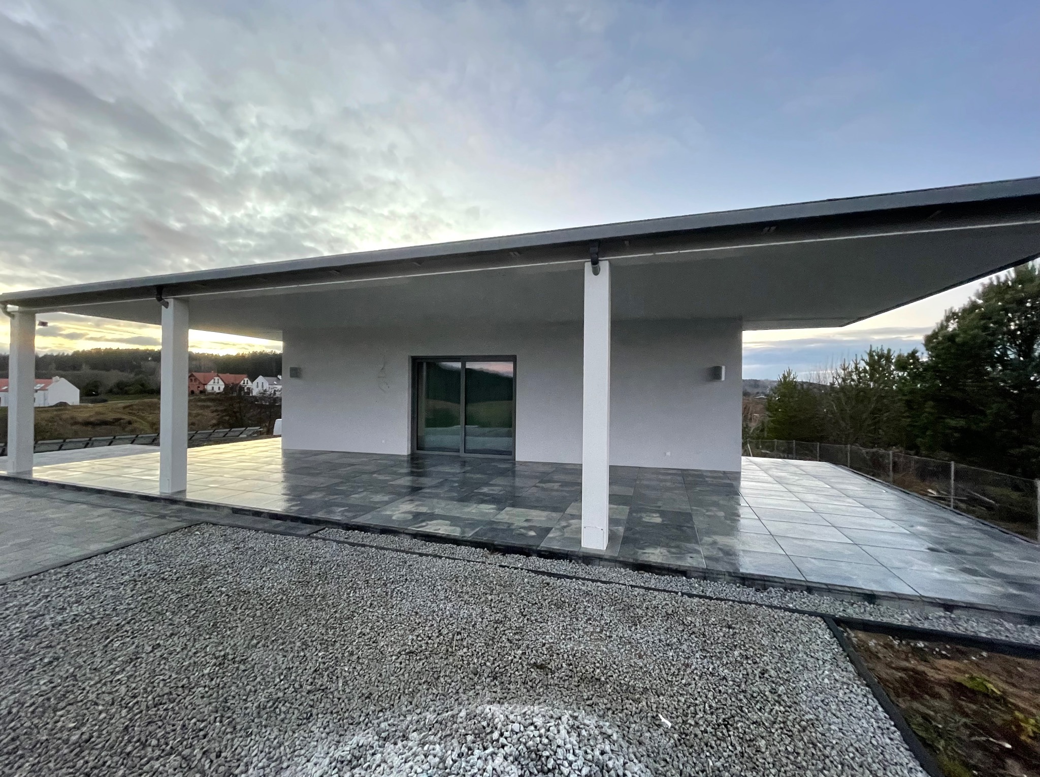 Nowoczesny taras z szarych płyt, wykończony drobnym żwirem. Minimalistyczna architektura budynku w tle, pochmurne niebo. Widok z poziomu gruntu.