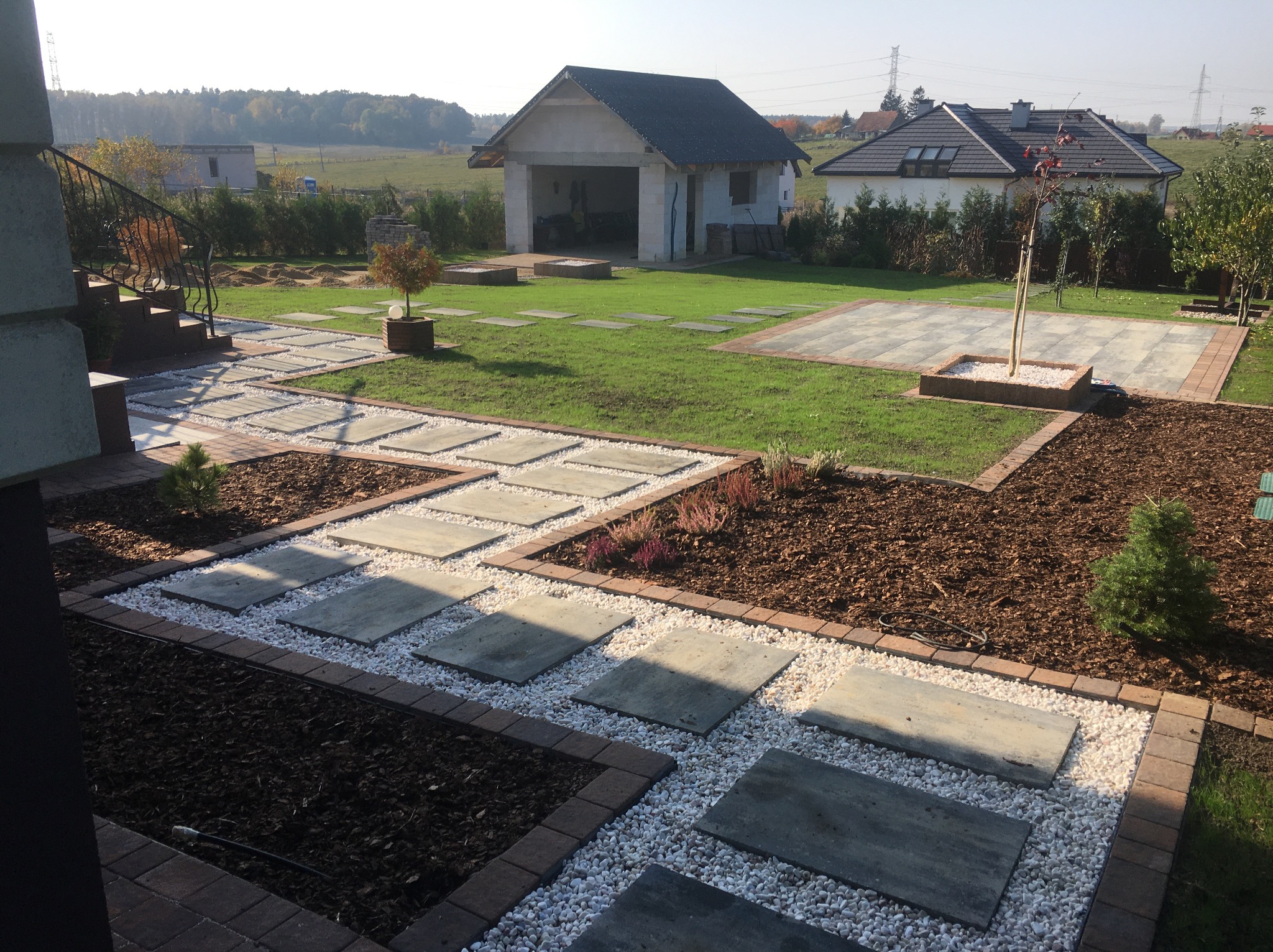 Ogród z ścieżką z płyt betonowych i białego żwiru, obrzeża z kostki, trawnik, budynek gospodarczy w tle, słoneczny dzień.