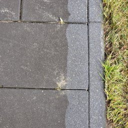 Brukodnowa - Częściowo umyta, szara kostka brukowa sąsiadująca z zieloną trawą. Widoczna różnica w czystości powierzchni kostki.