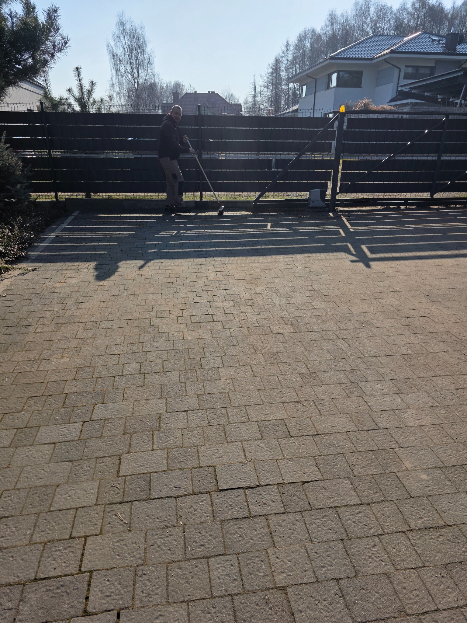 Pracownik zamiata kostkę brukową przed posesją z bramą wjazdową i domem w tle. Widoczne cienie, słoneczny dzień. Kostka o nieregularnej fakturze.