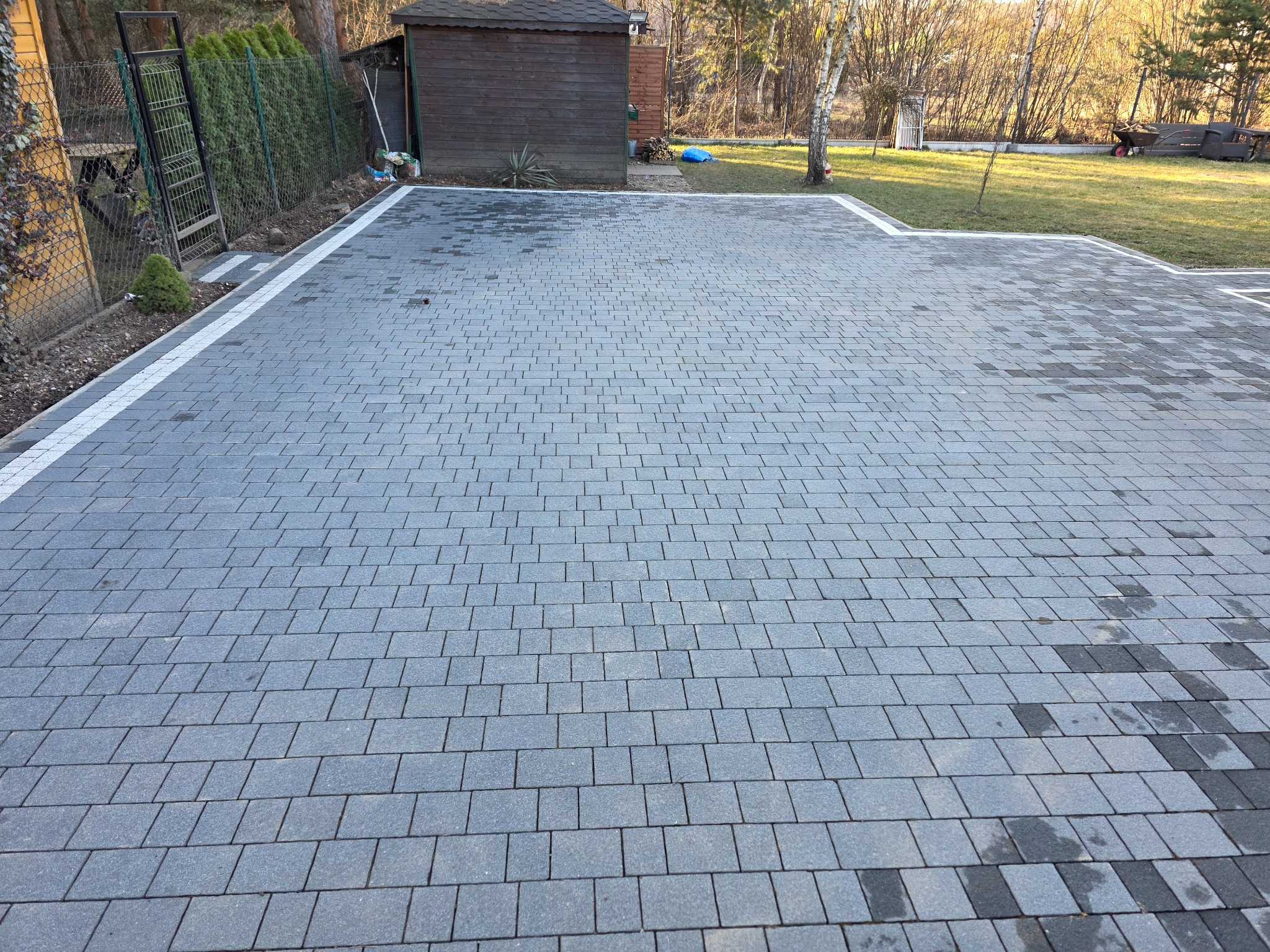 Wyłożona szara kostka brukowa z białą obwódką na posesji w Siemianowicach Śląskich. Widoczny fragment trawnika, budynek gospodarczy i ogrodzenie.