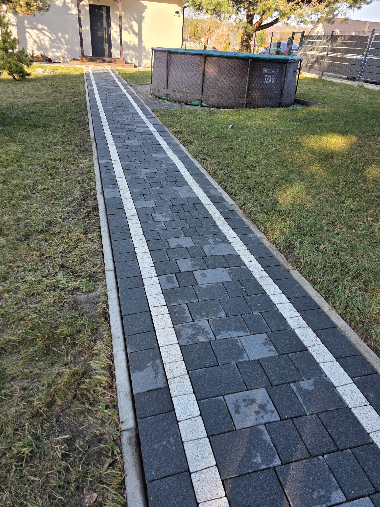 Ścieżka z ciemnej kostki brukowej z białymi pasami, prowadząca do domu z basenem w tle. Widok z perspektywy, ukazujący detale kostki i otoczenie.