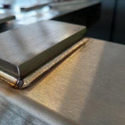 MaxPol - Precyzyjne łączenie metalowych elementów widoczne z bliska, ukazujące estetyczne spawy na powierzchni obrabianego metalu. Detal spoiny z efektem tęczy.