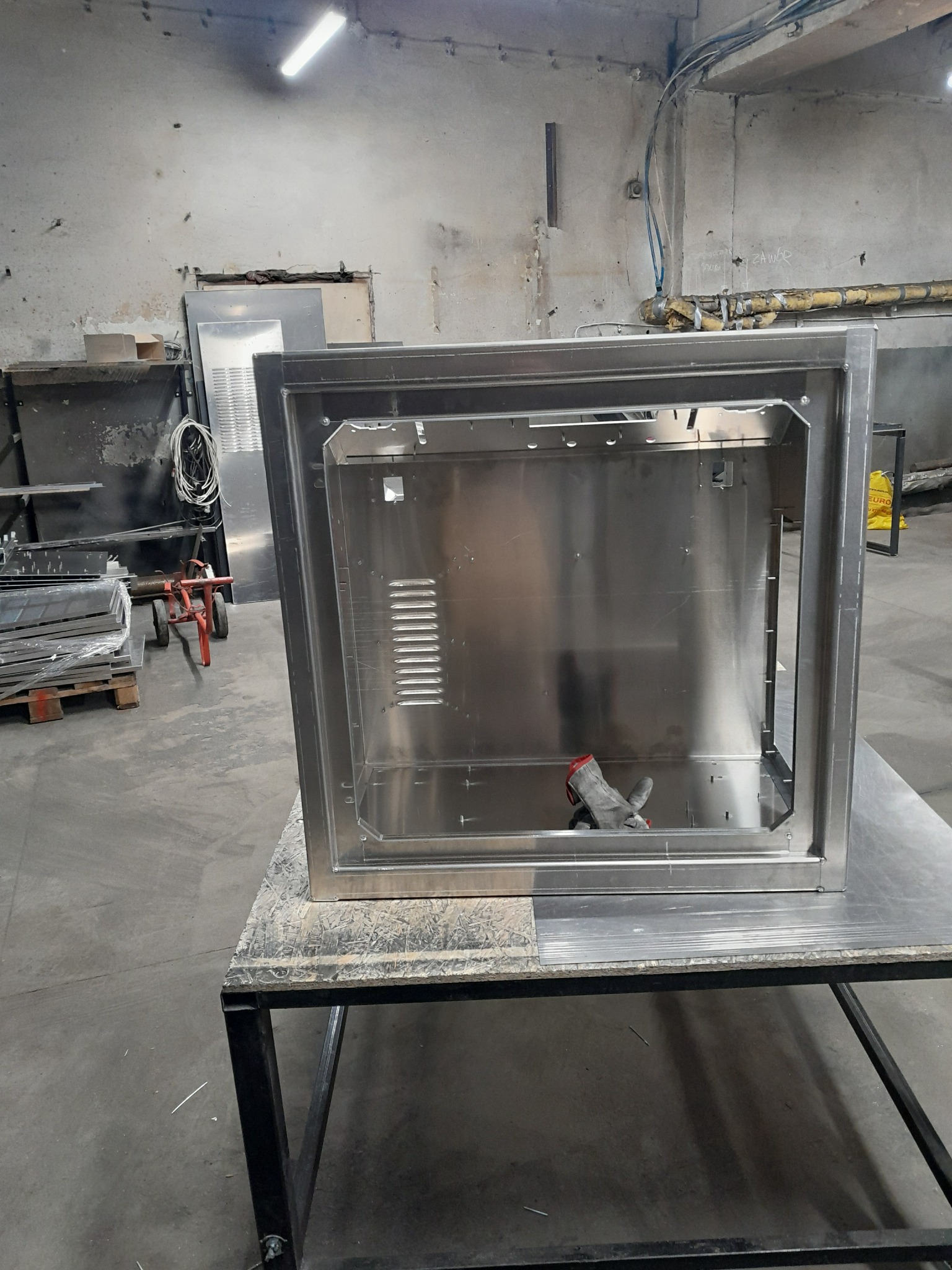 Spawana, metalowa obudowa urządzenia na stole warsztatowym. Widoczne ślady obróbki, spawania. W tle elementy warsztatu i składowane blachy.