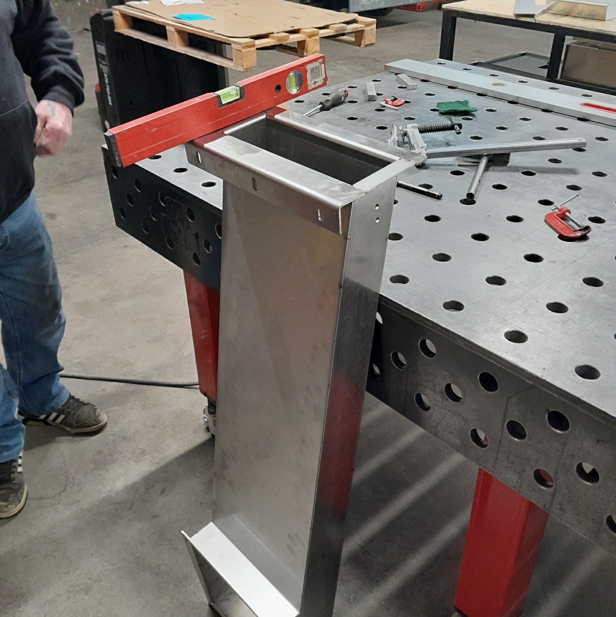 Metalowa konstrukcja na perforowanym stole warsztatowym, poziomica, narzędzia. Elementy zespawane, widoczne spawy. Częściowo widoczna osoba w roboczym ubraniu.