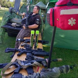 IVAN-OLEKSANDR Yakymiv Centrum Szkoleń - Mężczyzna w stroju medyka siedzi za stołem z replikami broni ASG, obok torba medyczna i imitacja wyrzutni rakiet. Scena z imprezy airsoftowej.