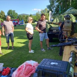 IVAN-OLEKSANDR Yakymiv Centrum Szkoleń - Pokaz sprzętu militarnego na imprezie plenerowej: karabiny, granatniki, kamizelka taktyczna. W tle dmuchany zamek i publiczność.