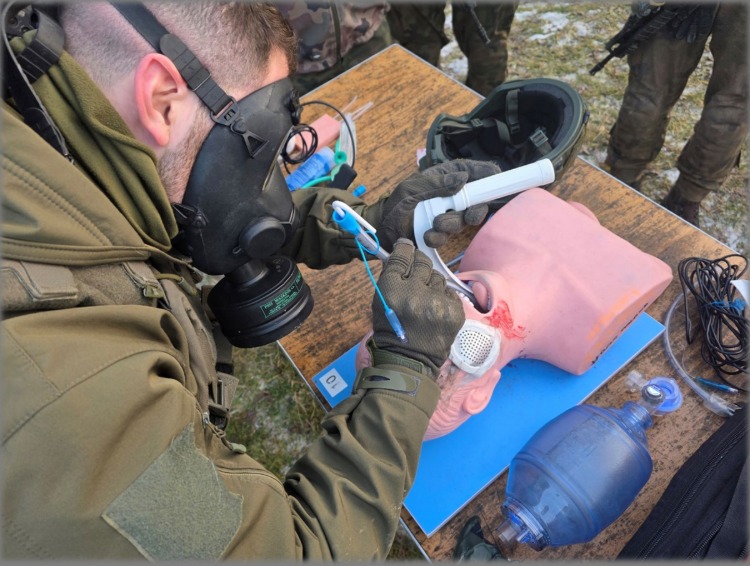 Instruktor w masce gazowej demonstruje intubację na fantomie medycznym podczas kursu pierwszej pomocy w warunkach poligonowych. Widoczny sprzęt medyczny i umundurowani uczestnicy.