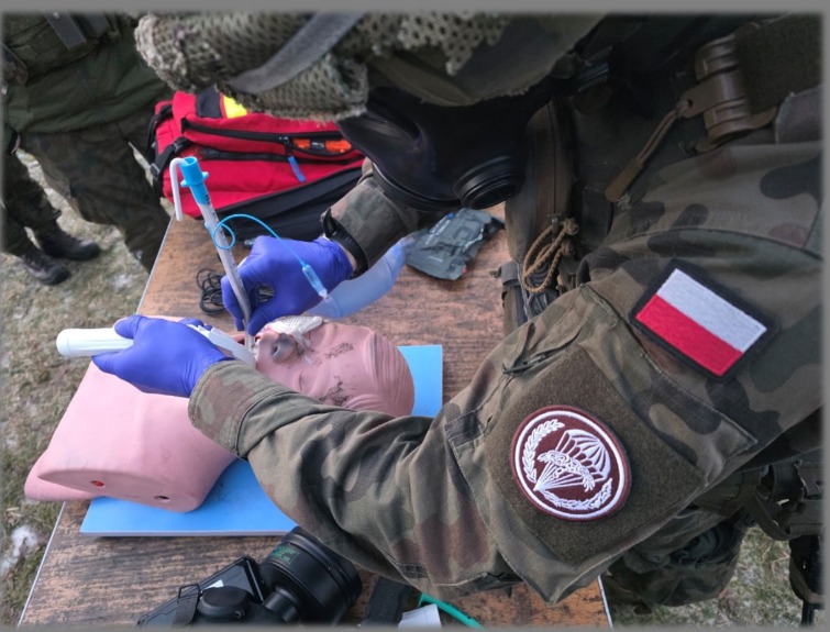 Instruktor w mundurze z flagą Polski i maską gazową demonstruje intubację na manekinie podczas kursu pierwszej pomocy. Widoczny sprzęt medyczny i otoczenie poligonowe.