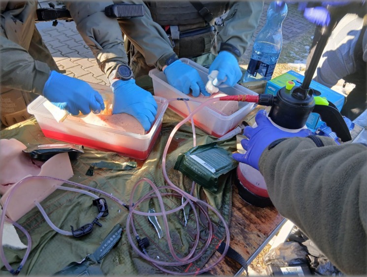 Symulacja medyczna: instruktorzy w rękawicach demonstrują techniki ratunkowe na manekinach z krwią, używając pompki i opatrunków na stole podczas kursu pierwszej pomocy.