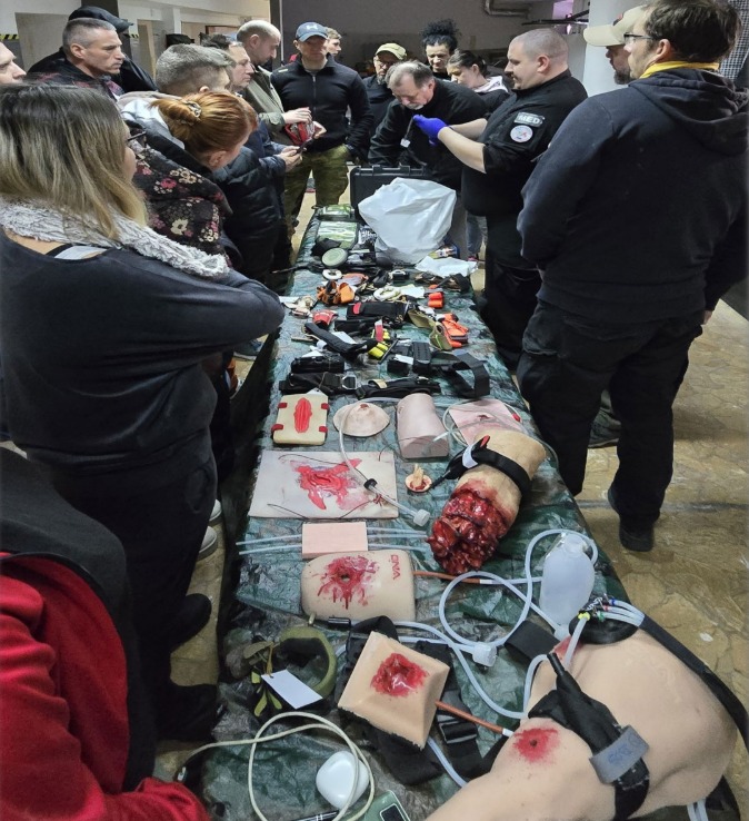 Kurs pierwszej pomocy: grupa osób obserwuje instruktora demonstrującego techniki ratunkowe na manekinach z symulowanymi ranami i sprzętem medycznym na stole.