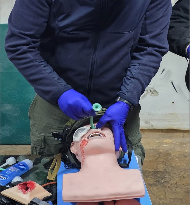 Instruktor w niebieskich rękawicach demonstruje intubację na manekinie z symulacją ran podczas kursu pierwszej pomocy. Widoczne akcesoria medyczne.