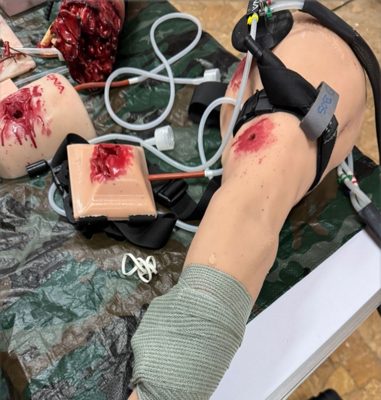 Manekiny z symulowanymi ranami, bandaże i akcesoria medyczne na tle moro, używane podczas kursu pierwszej pomocy, prezentujące realistyczne scenariusze.