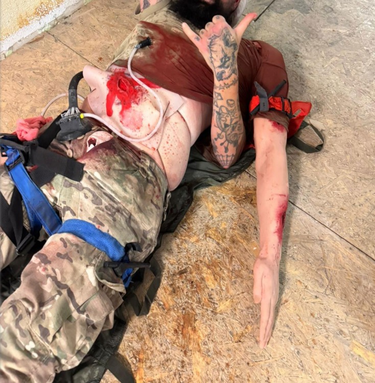 Manekin z symulowanymi ranami i założonymi opatrunkami, używany podczas szkolenia z pierwszej pomocy. Widoczne realistyczne obrażenia i sprzęt medyczny.