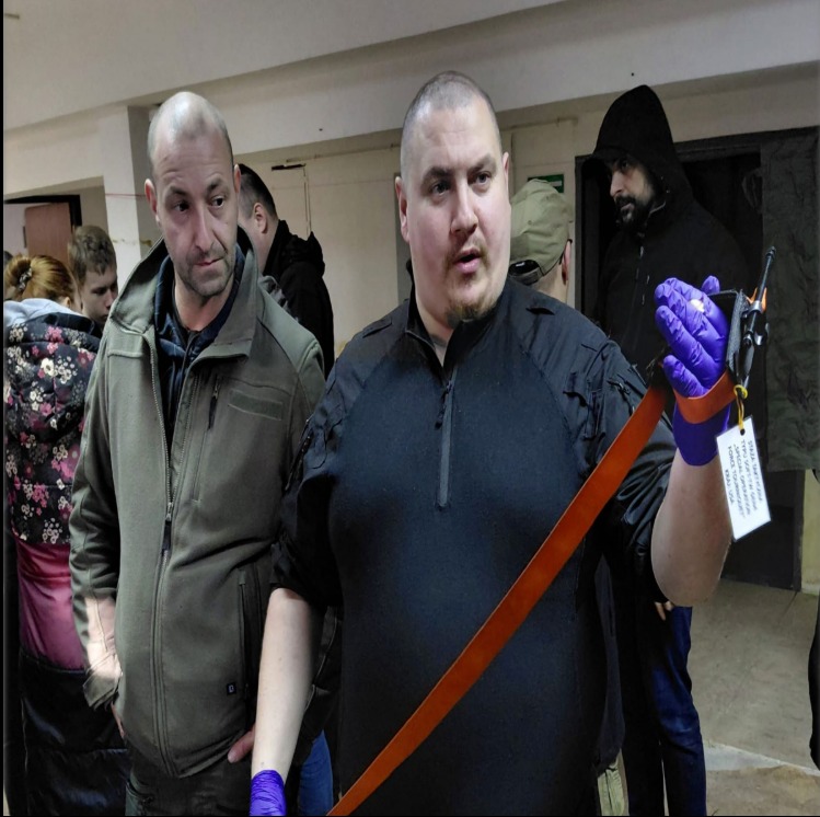 Instruktor w rękawiczkach prezentuje stazę taktyczną na tle grupy osób podczas szkolenia z pierwszej pomocy. Widoczny kursant obserwujący demonstrację.