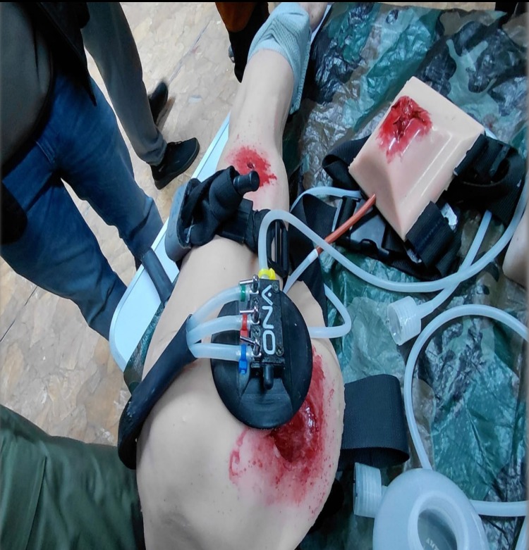 Manekin nogi z symulacją rany i systemem do ćwiczeń tamowania krwotoku, używany podczas kursu pierwszej pomocy. Widoczne elementy wyposażenia i osoby w tle.