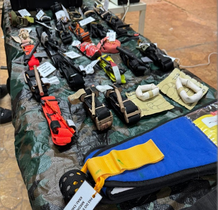 Różnorodny sprzęt medyczny, w tym stazy taktyczne i bandaże, rozłożony na stole z wzorzystym obrusem. Widoczne etykiety i opakowania. Sprzęt do udzielania pierwszej pomocy.