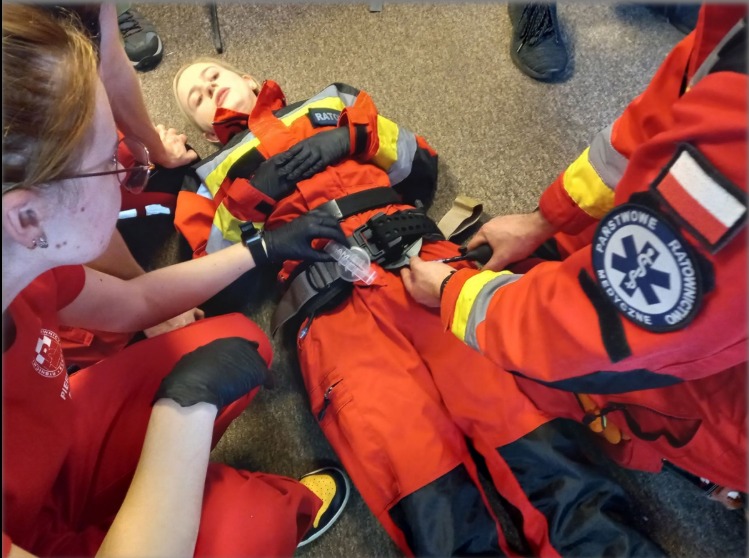 Symulacja udzielania pierwszej pomocy poszkodowanemu przez ratowników medycznych w czerwonych uniformach. Widoczny manekin i sprzęt medyczny.