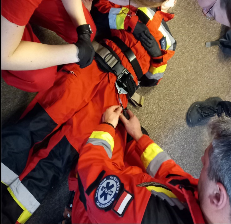 Przygotowanie do symulacji ratowniczej: instruktorzy zapinają pasy i sprawdzają sprzęt na manekinie ubranym w strój ratownika medycznego. Ćwiczenia praktyczne.