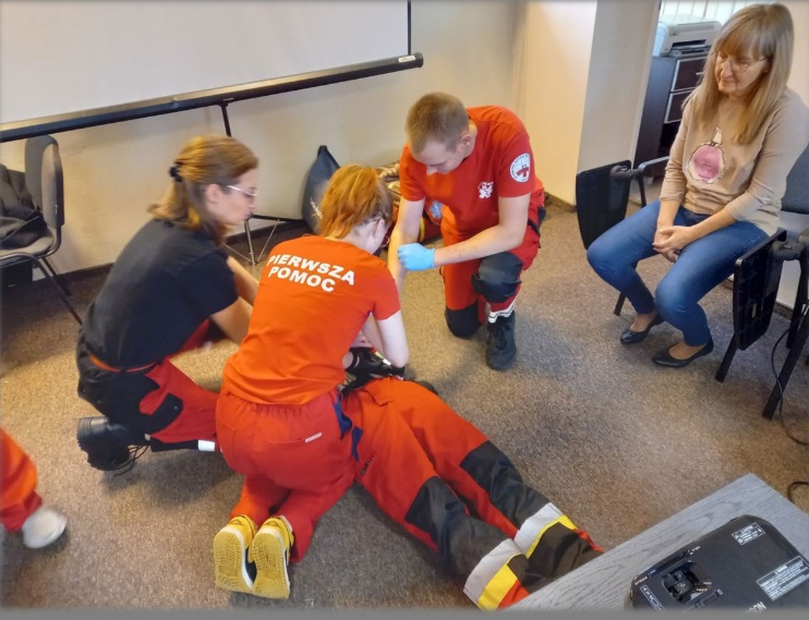 Instruktorzy w czerwonych uniformach demonstrują resuscytację na manekinie podczas kursu pierwszej pomocy. Uczestniczka obserwuje z boku.