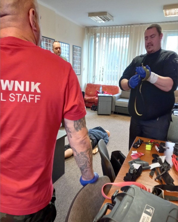 Instruktor zakłada stazę taktyczną na fantom podczas kursu pierwszej pomocy. Widoczny sprzęt medyczny i drugi instruktor w czerwonej koszulce.