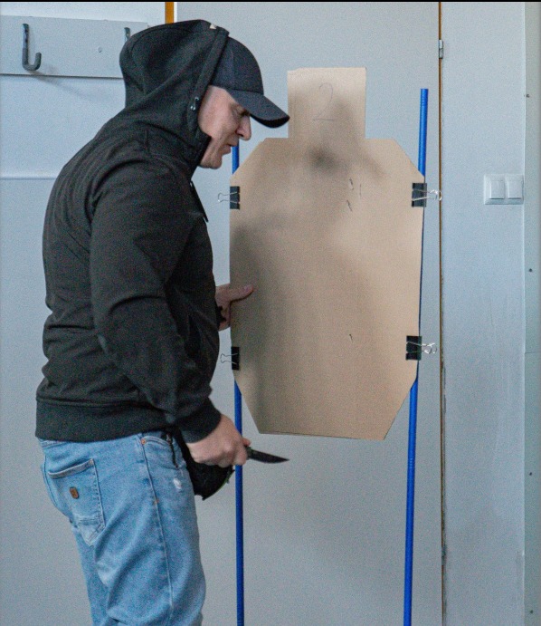 Mężczyzna w bluzie z kapturem i czapce, trzymający nóż, stoi przed kartonową sylwetką człowieka przymocowaną do niebieskich rurek. Ćwiczenia z zakresu samoobrony.