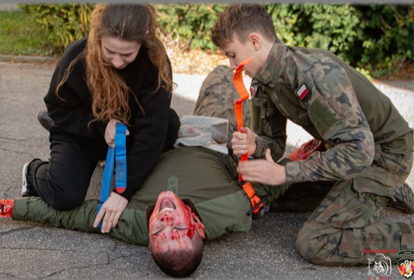 Symulacja udzielania pierwszej pomocy poszkodowanemu z ranami głowy. Osoby w trakcie szkolenia, jedna w cywilnym ubraniu, druga w mundurze wojskowym.