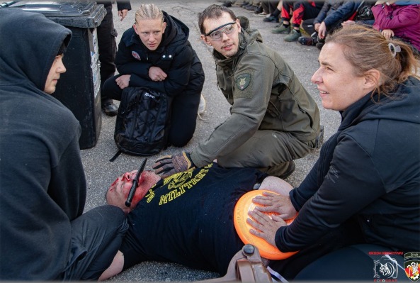 Symulacja udzielania pierwszej pomocy poszkodowanemu z atrapą noża w twarzy. Uczestnicy kursu w trakcie ćwiczeń praktycznych, widoczny tłum osób w tle.