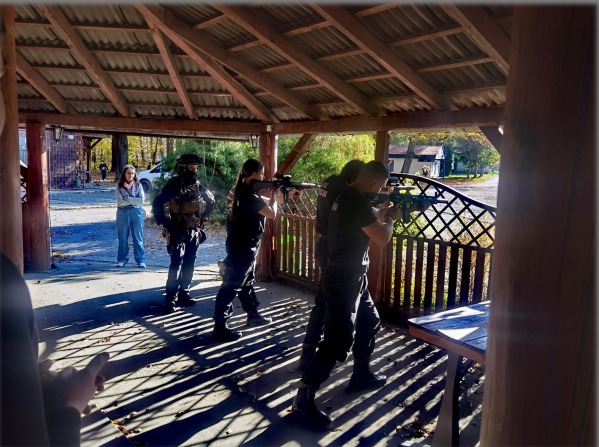 Szkolenie strzeleckie: grupa osób w czarnych strojach ćwiczy celowanie z broni pod drewnianą wiatą, z widoczną osobą obserwującą w tle.