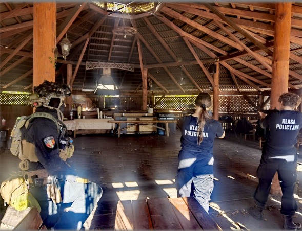 Zajęcia z taktyki i technik interwencji dla klasy policyjnej w plenerze. Uczestnicy w umundurowaniu i z replikami broni ćwiczą pod drewnianą wiatą.